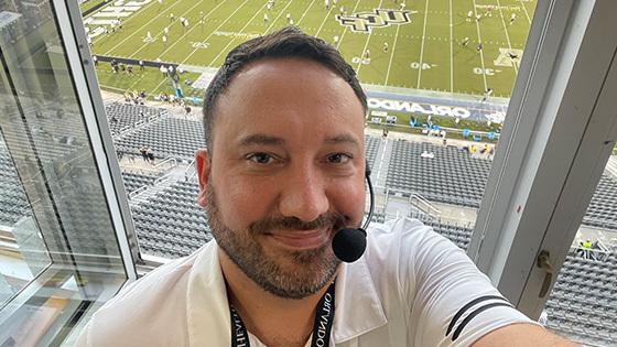 Jeff Sharon wears a headset microphone and smiles at the camera. He is in an announcer’s booth with the UCF football stadium below him.